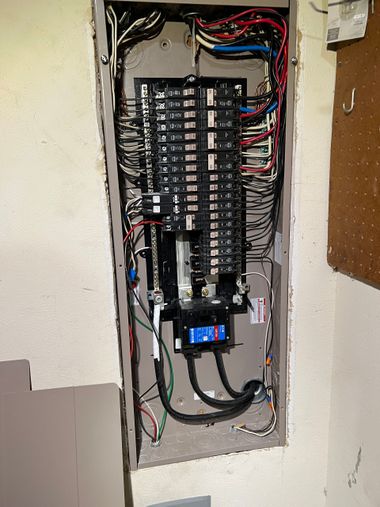 Open electrical breaker panel with exposed wiring and a digital meter in a wall recess