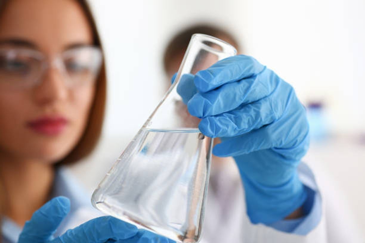 Scientist wearing blue gloves examines a transparent liquid in a conical flask, inside a lab. Scientist wearing blue gloves examines a transparent liquid in a conical flask, inside a lab.