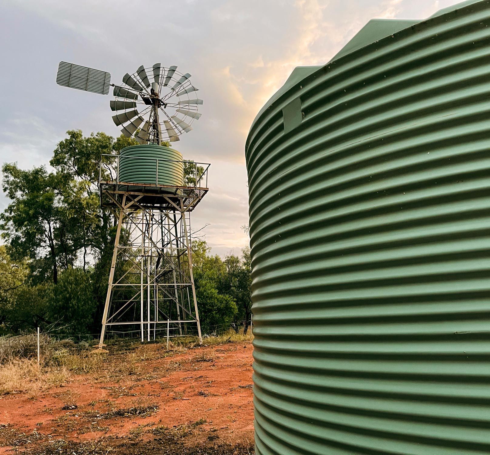 Windmill and water tank on reddish soil, with trees and a cloudy sky. Windmill and water tank on reddish soil, with trees and a cloudy sky.