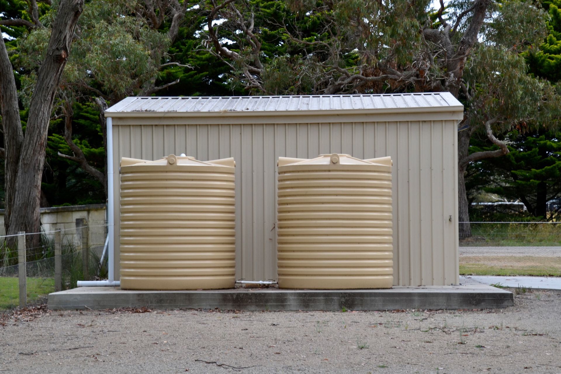 Two beige water tanks in front of a tan shed with a silver roof, set on a concrete pad. Two beige water tanks in front of a tan shed with a silver roof, set on a concrete pad.