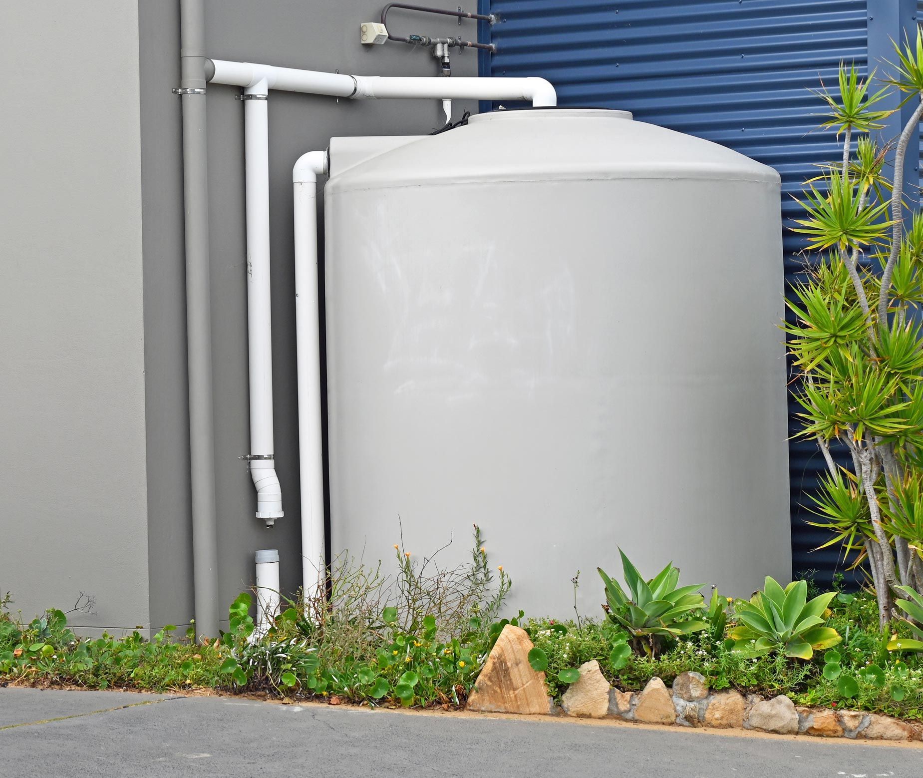 A large, grey water tank against a wall, with white pipes connected; surrounded by plants and a paved area. A large, grey water tank against a wall, with white pipes connected; surrounded by plants and a paved area.
