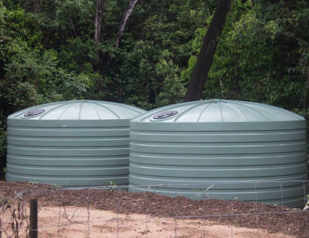 Two green, cylindrical water tanks with domed lids sit in a wooded area. Two green, cylindrical water tanks with domed lids sit in a wooded area.