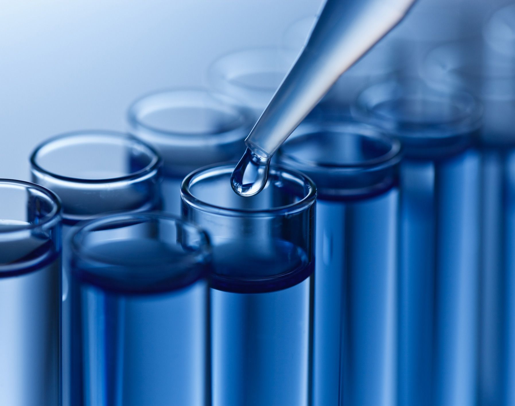 A dropper dispensing liquid into a test tube with other test tubes in the background, all filled with blue liquid. A dropper dispensing liquid into a test tube with other test tubes in the background, all filled with blue liquid.
