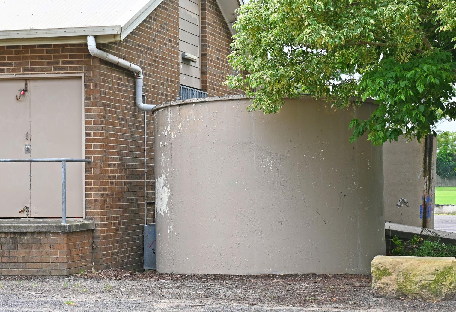 A tan cylindrical water tank next to a brick building, with a downspout and tree visible. A tan cylindrical water tank next to a brick building, with a downspout and tree visible.