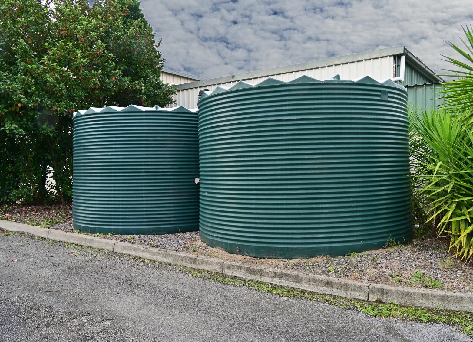 Two green corrugated metal water tanks on a concrete pad, surrounded by greenery and a building in the background. Two green corrugated metal water tanks on a concrete pad, surrounded by greenery and a building in the background.
