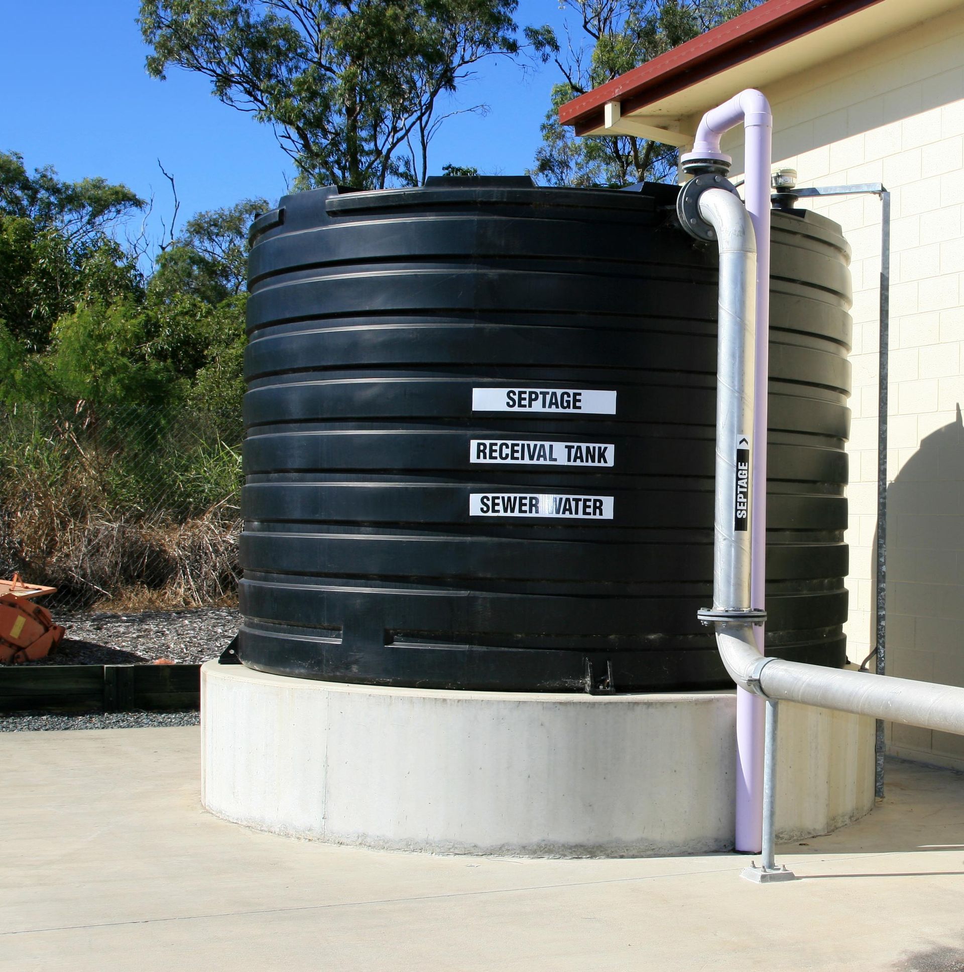 Black septic tank labeled 'Septage', 'Recital Tank', 'Sewer Water', against a building, pipes attached. Black septic tank labeled 'Septage', 'Recital Tank', 'Sewer Water', against a building, pipes attached.