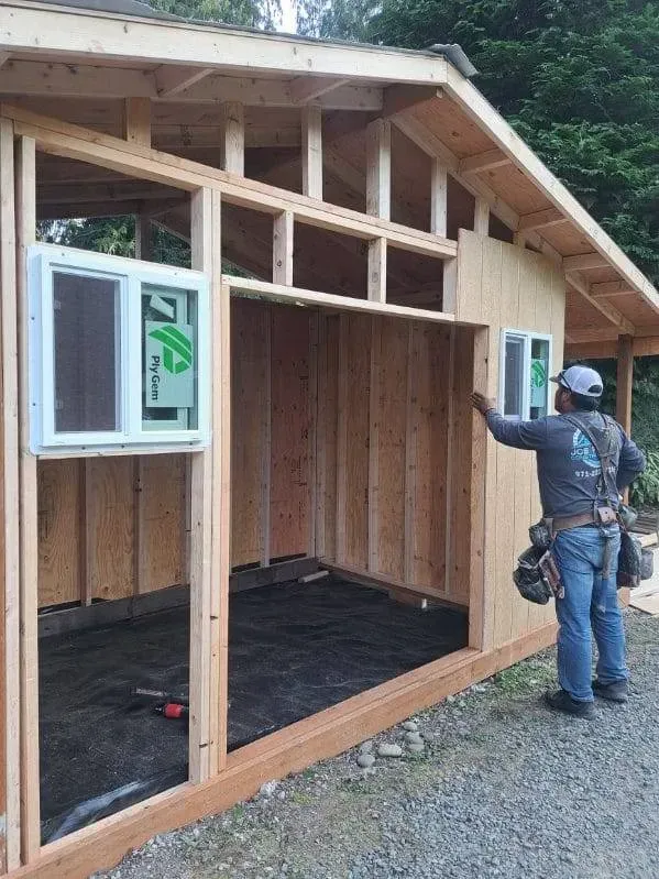 Person inspecting unfinished wooden shed with open framing and a window near the left side.