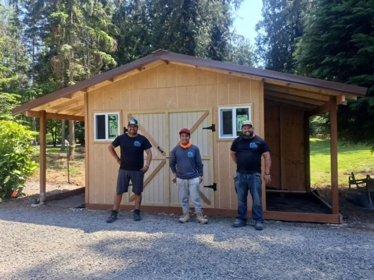 Three people standing outside a small wooden cabin with a porch, surrounded by trees.