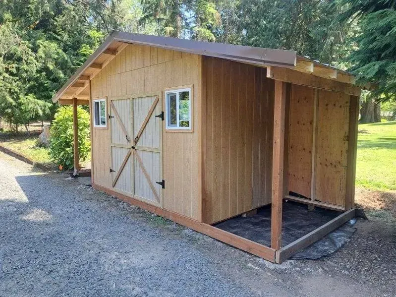 Wooden shed with double barn doors and open side, set on gravel beside a grassy wooded area.