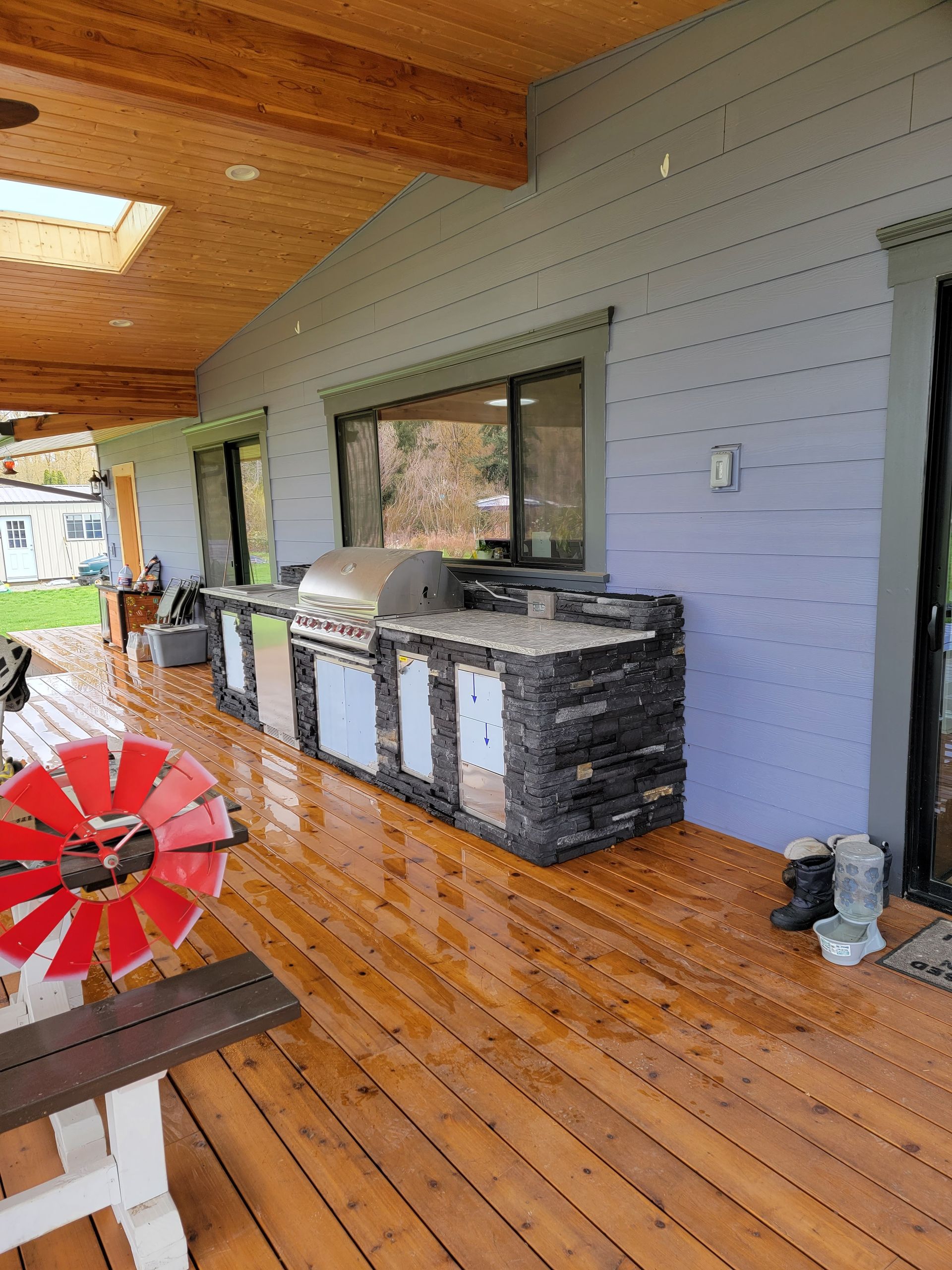 Covered porch with wooden floor, gray wall, windows, and a black outdoor grill or bar unit.