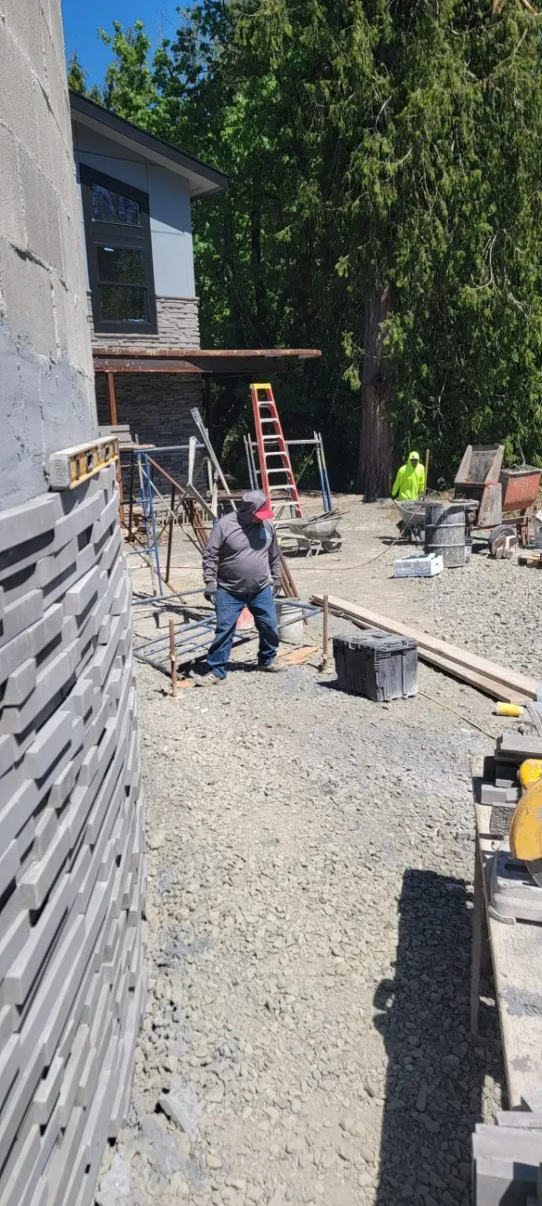 Workers on a gravel construction site beside a building, with ladders, tools, and stacked materials.