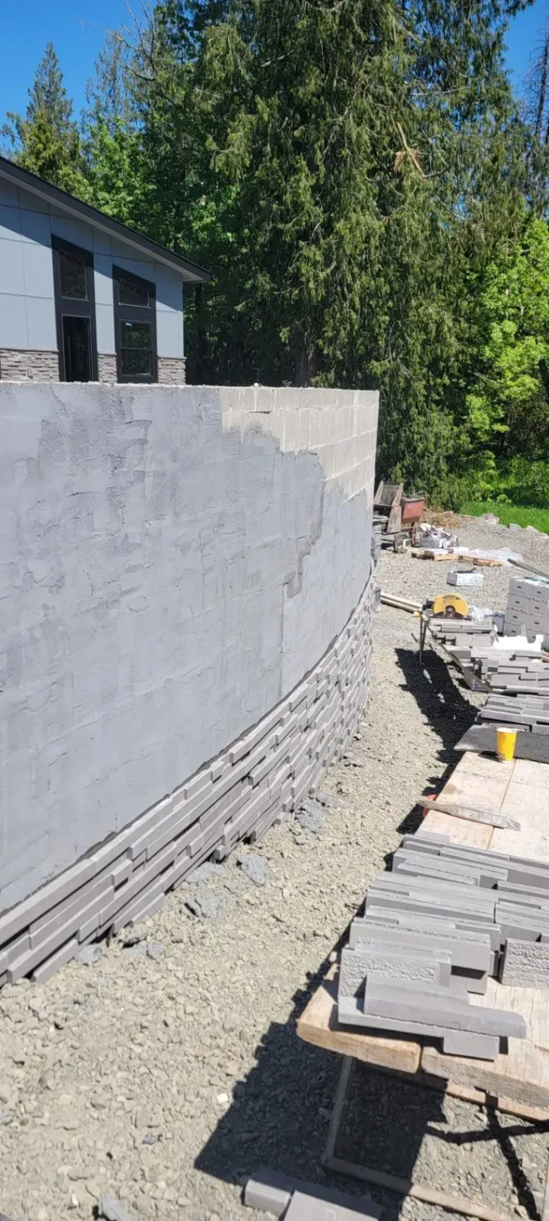 Curved concrete retaining wall under construction beside a house, with stacked pavers and gravel.