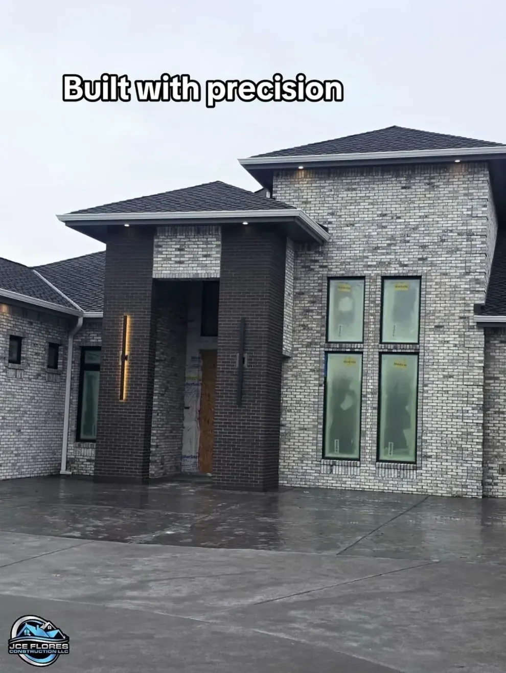 Modern gray stone house exterior with large windows and a paved driveway under an overcast sky