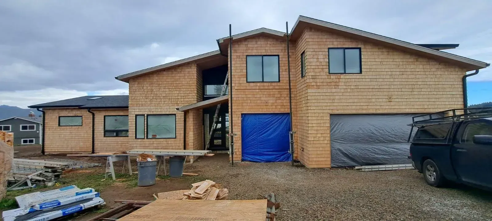 Two-story unfinished brick house with garage doors, blue tarp, and construction materials on a gravel lot