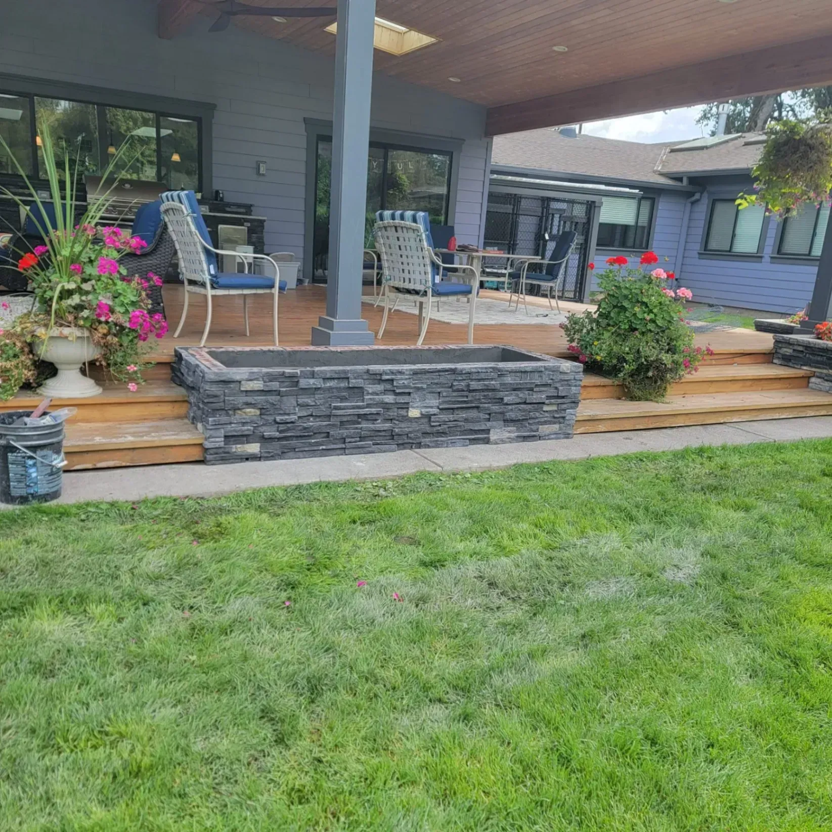 Front porch with stone retaining wall, steps, patio chairs, hanging flowers, and a green lawn.
