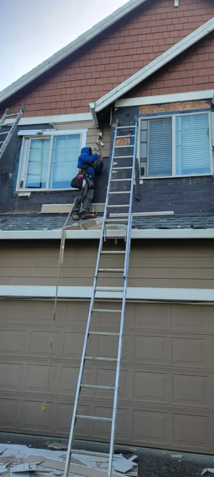 Worker on a tall ladder repairing the roof edge of a house above a garage.
