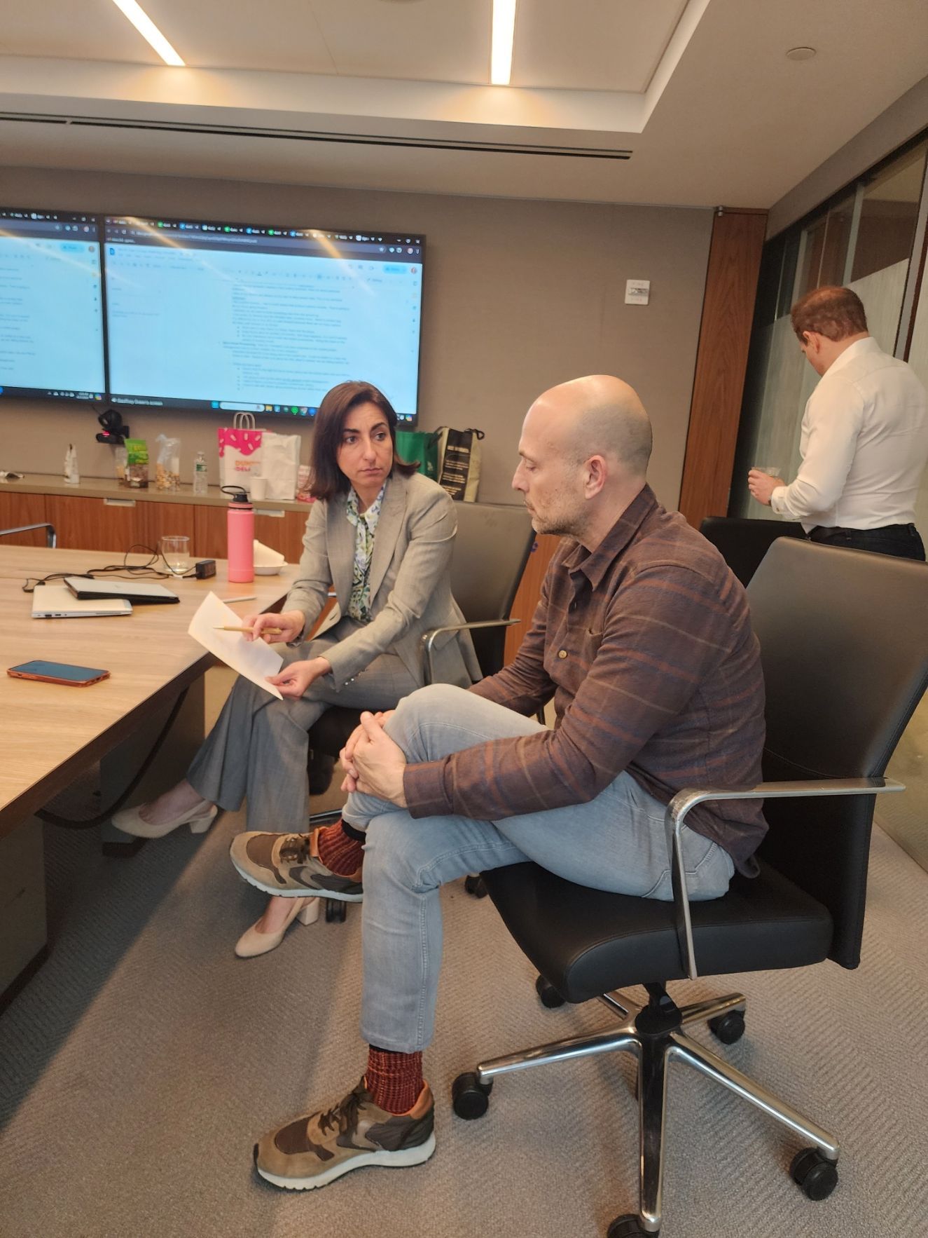 A woman points at a glass wall covered with sticky notes during a meeting. Others observe. A woman points at a glass wall covered with sticky notes during a meeting. Others observe.