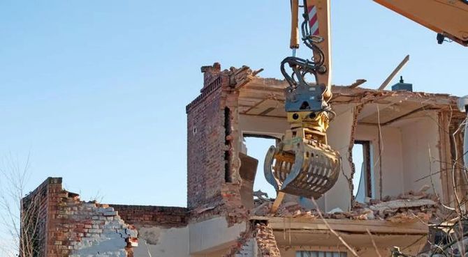 Excavator demolishing a partially collapsed brick building under a clear blue sky