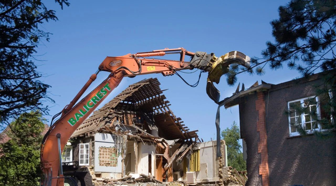 Excavator demolishing a partially collapsed house beside a brick building under a clear blue sky
