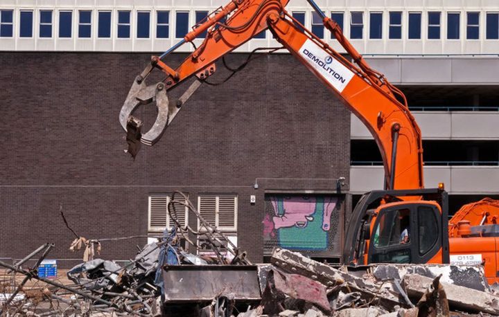 Orange excavator demolishing rubble beside a dark wall on a construction site