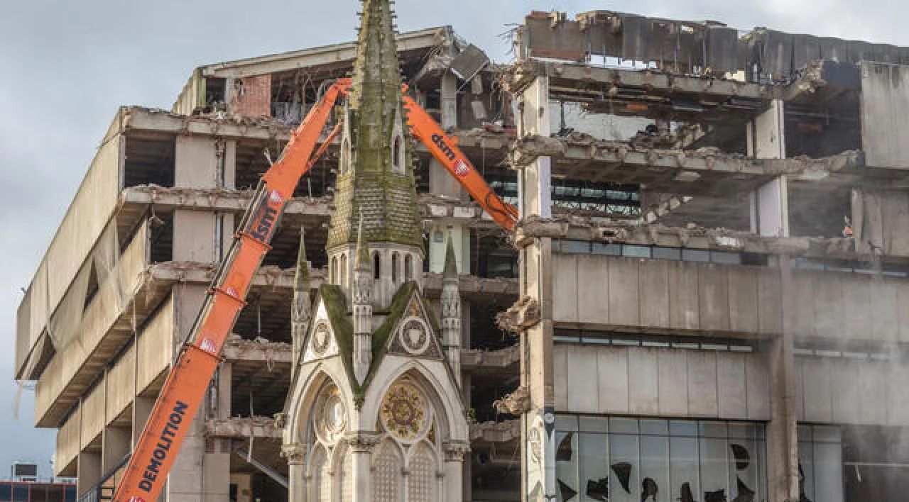 Church steeple beside a damaged building with orange cranes during demolition or repair