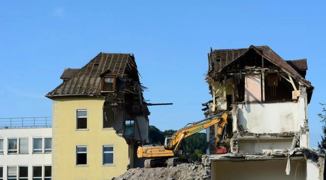 Demolition excavator tearing down a damaged two-story house next to an intact yellow building.