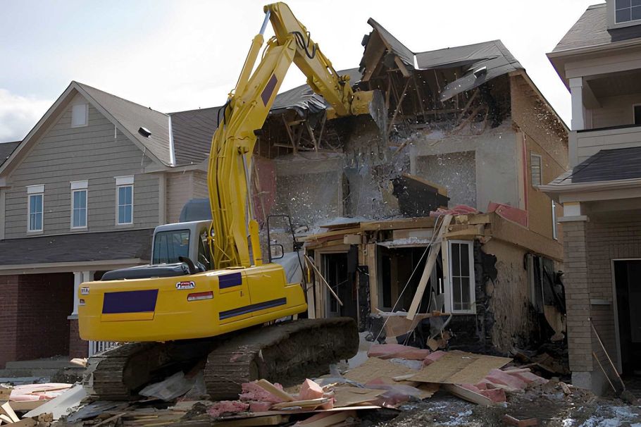Yellow excavator demolishing the front of a two-story house, debris scattered across the yard.