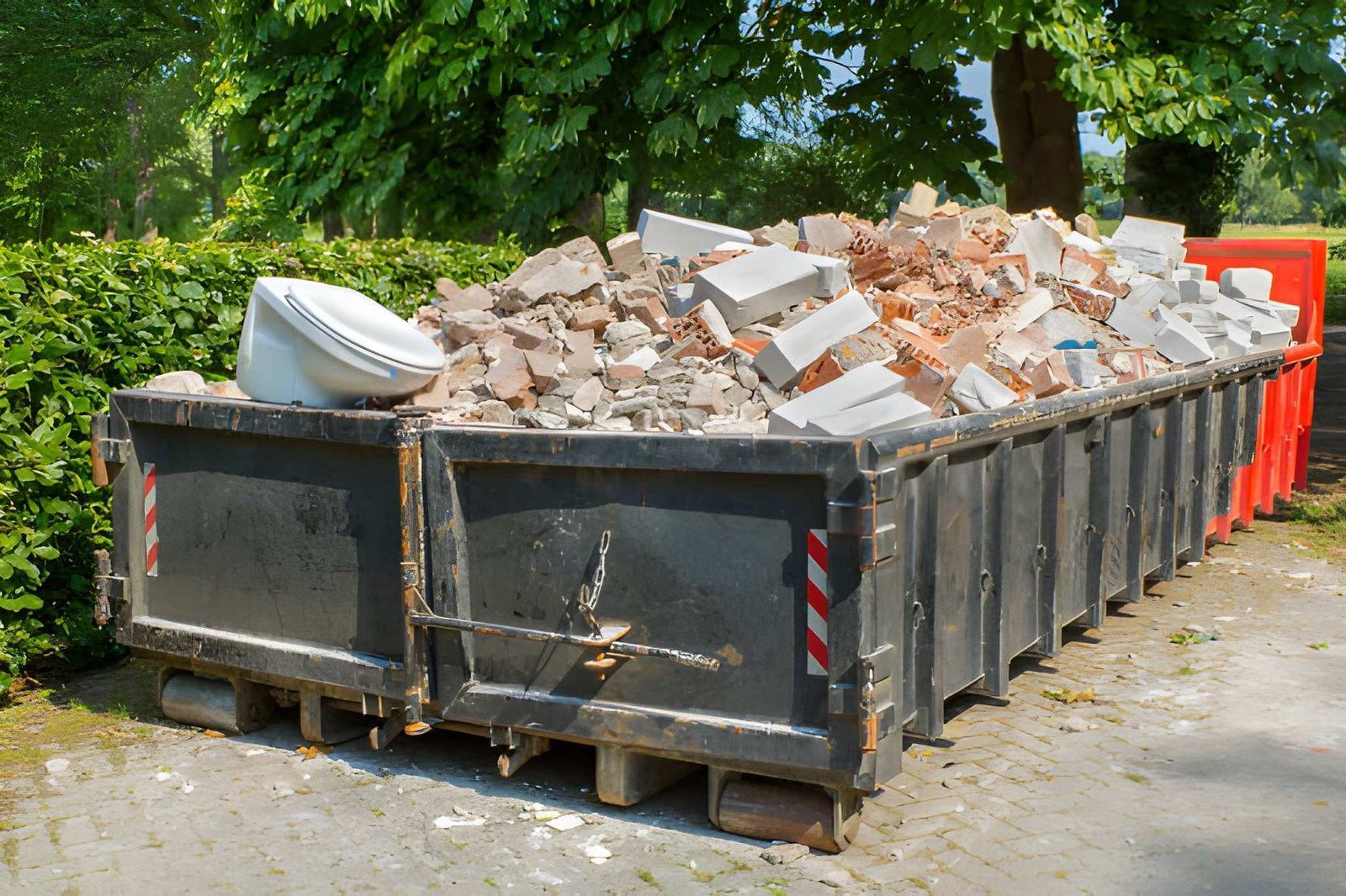Construction dumpster full of rubble and bricks on a gravel road, with trees in the background