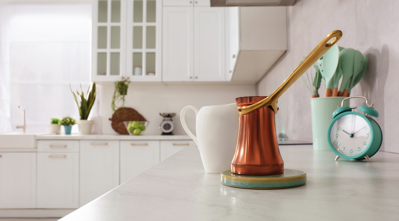 A kitchen counter with a coffee pot , cup , clock and utensils.