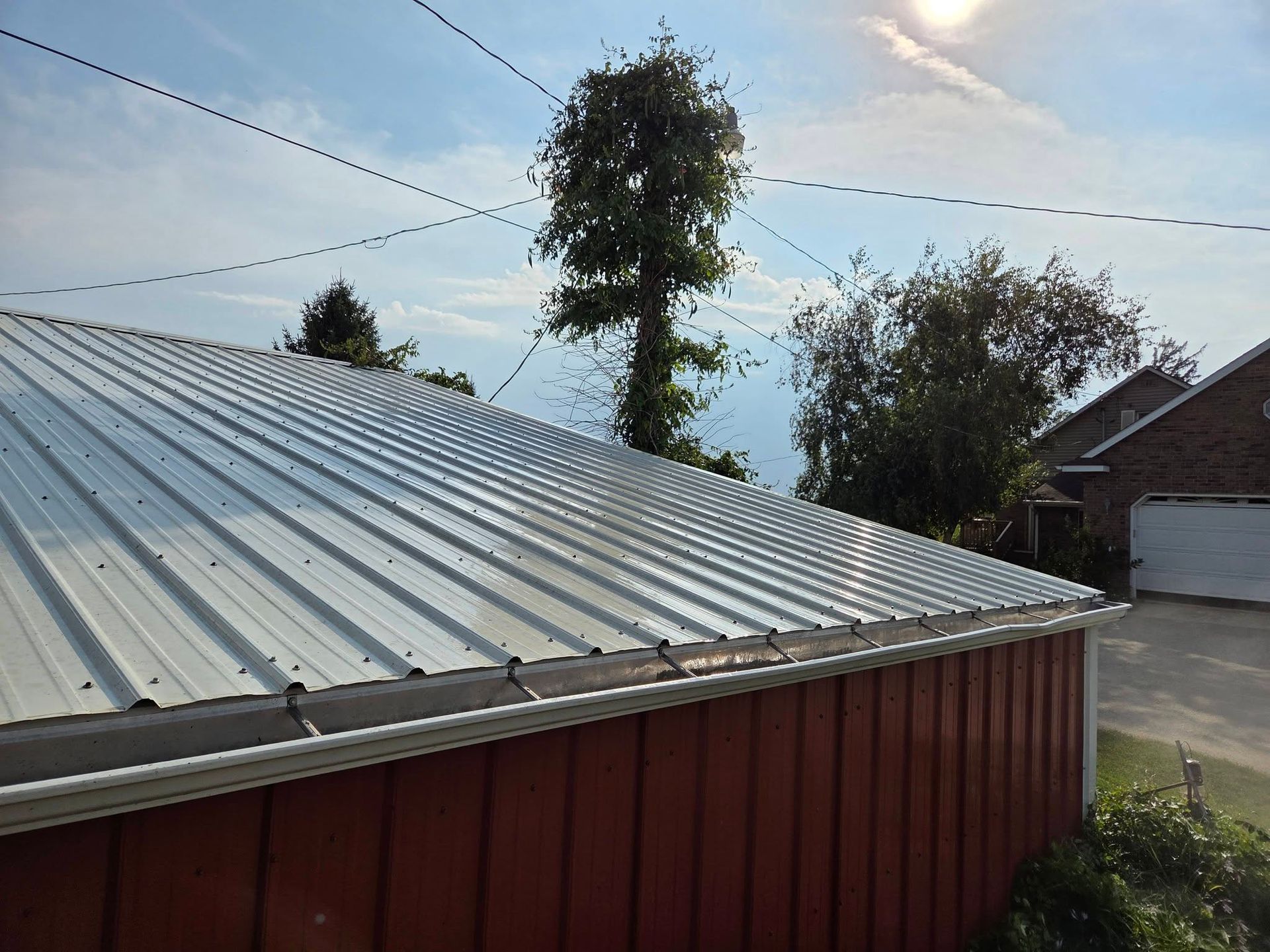 Red metal roof with silver ridges beside a road, trees, and a house under a partly cloudy sky.