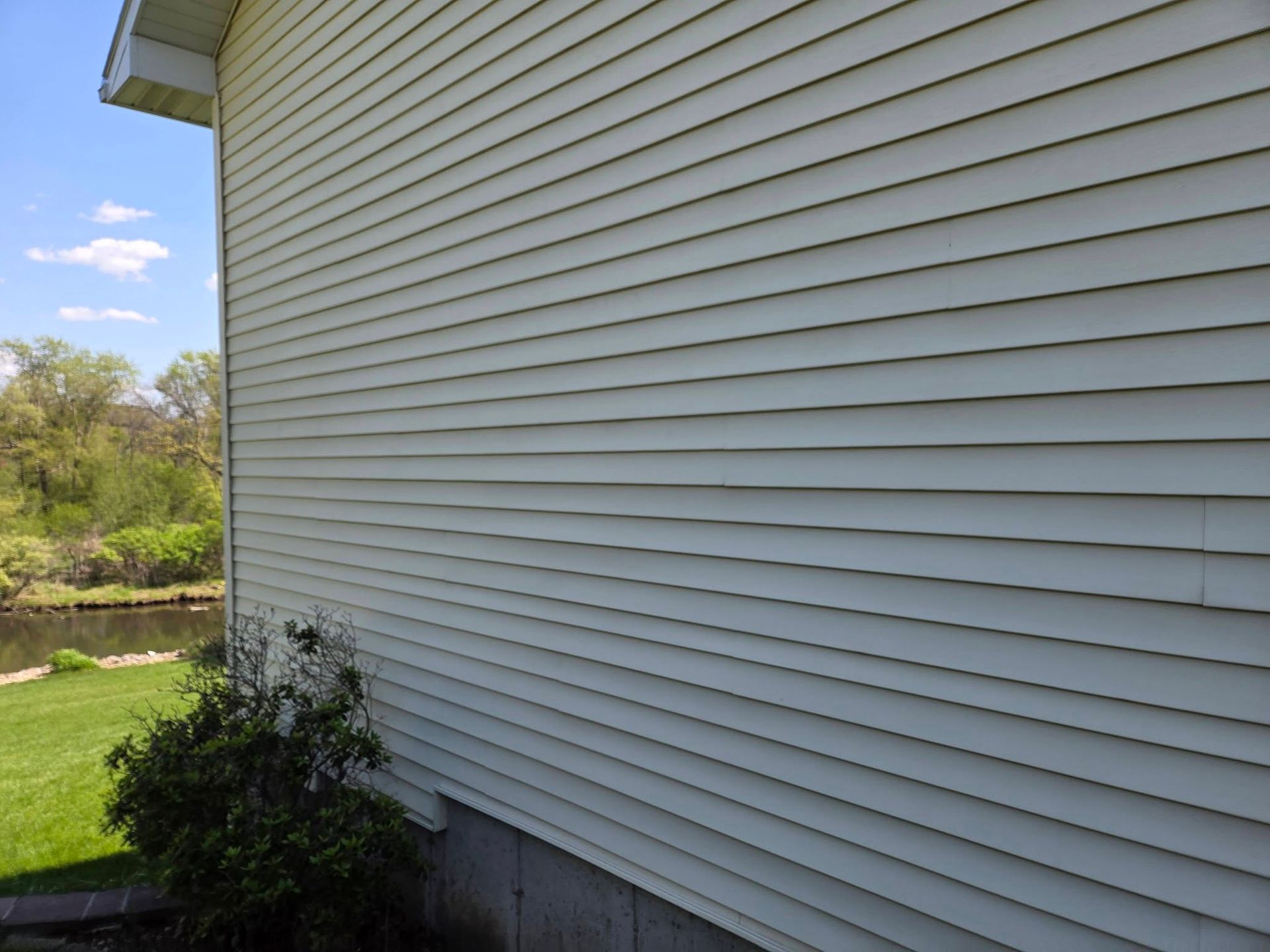 Side view of a white house with vinyl siding, a small shrub, and green lawn under a blue sky.