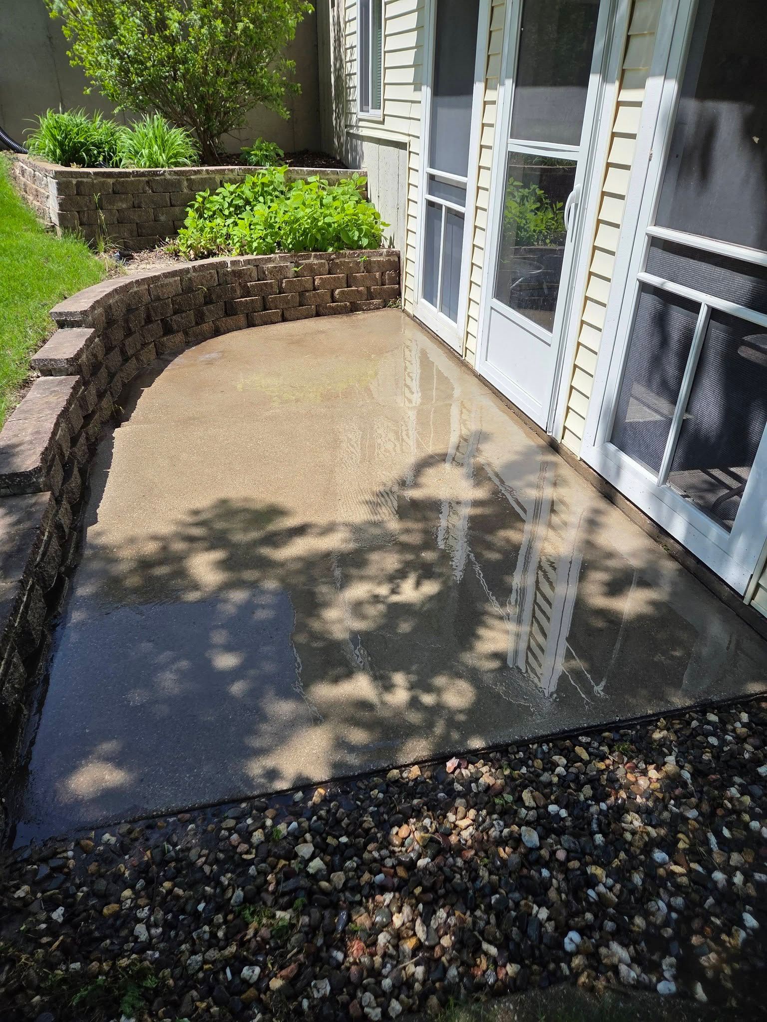 Curved stone patio beside a house with wet concrete and white-framed glass doors in sunlight