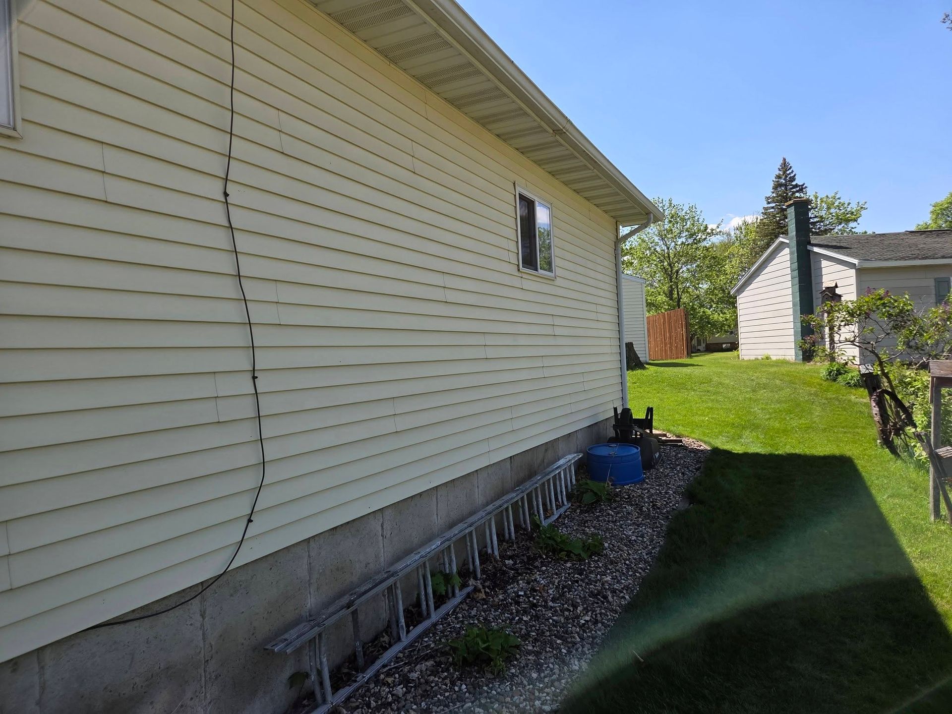 Side yard beside a white house with gravel bed, green lawn, and blue bin near the back