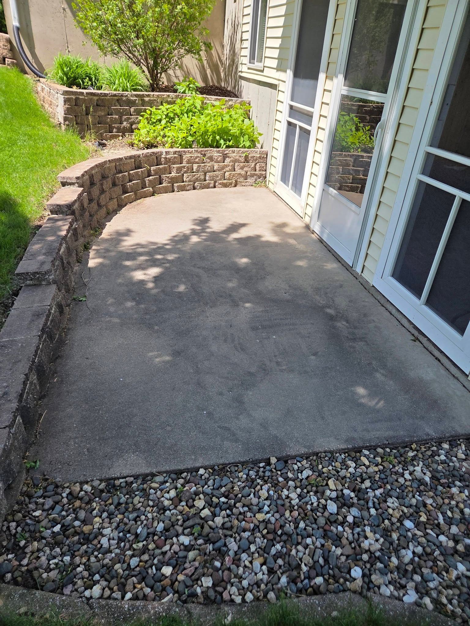 Small concrete patio by a glass door, bordered by stone retaining walls and gravel.