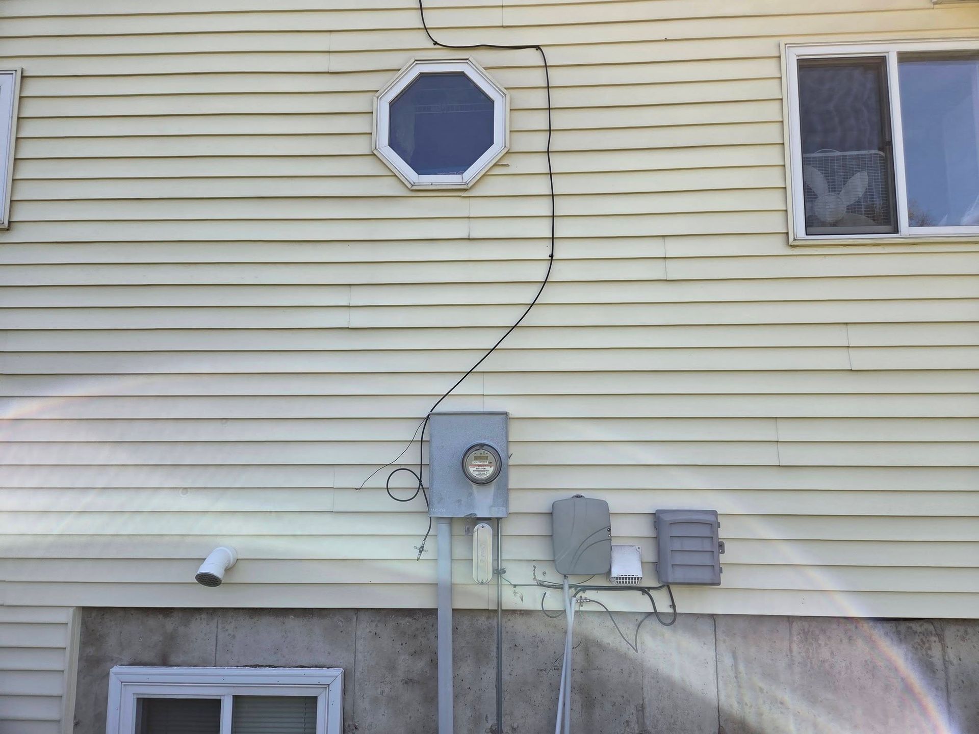 Side of a beige house with utility meters, cables, a small octagonal window, and one rectangular window