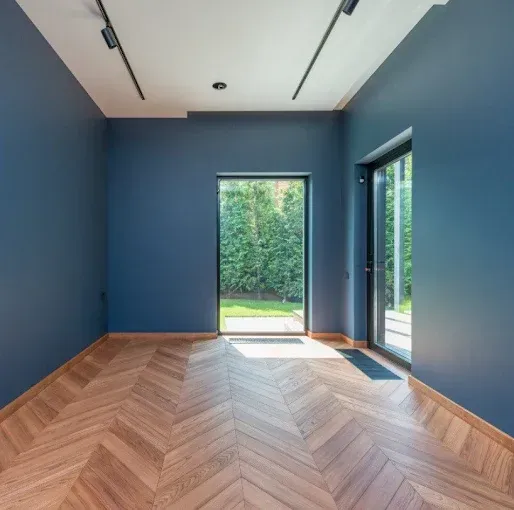Empty blue room with herringbone wood floor and large windows opening to greenery
