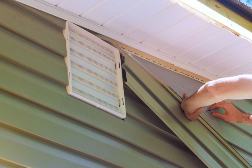 Hand reaching to adjust green exterior siding near a white vent under a roof eave