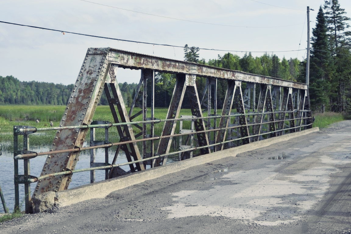 Metal truss bridge over water beside a gravel road in a rural landscape