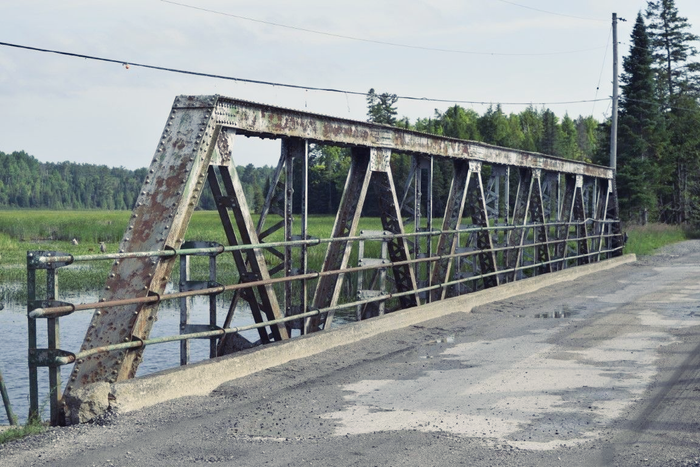 Metal truss bridge over water beside a gravel road in a rural landscape