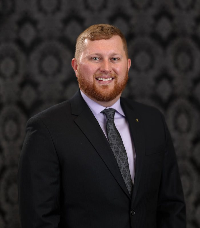 Man in a suit smiles at the camera, with a patterned dark background.
