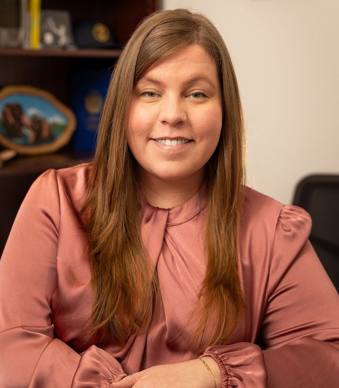 Woman with brown hair smiles, wearing a pink shirt, in an office setting.