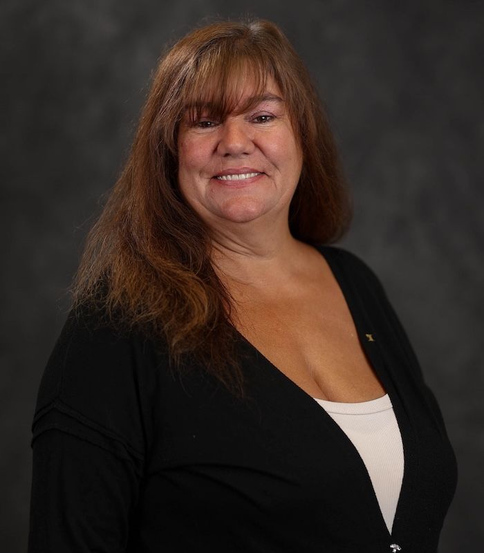 Woman with long brown hair, wearing a black cardigan over a white top, smiling in front of a gray backdrop.