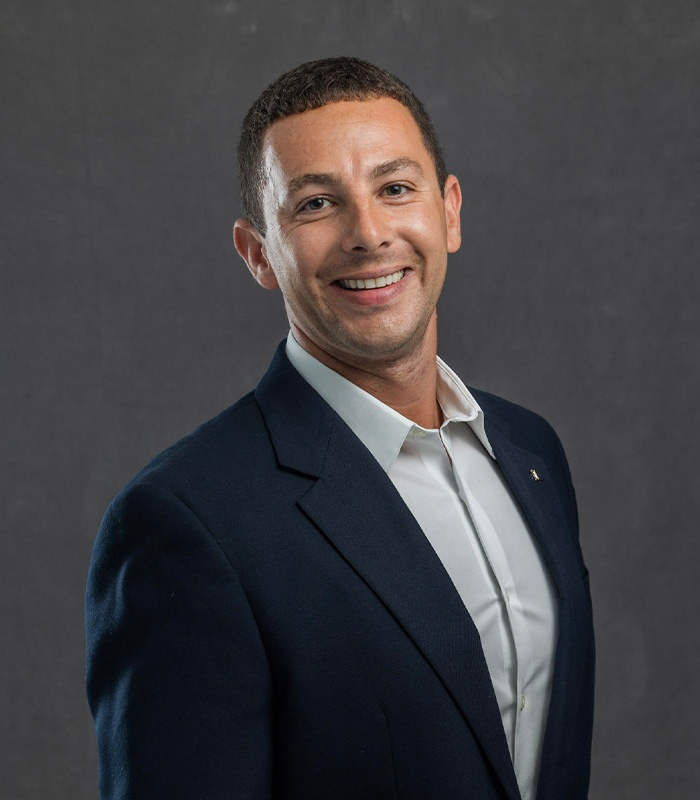 Man in navy blazer and white shirt smiles, looking towards the viewer. Dark gray background.
