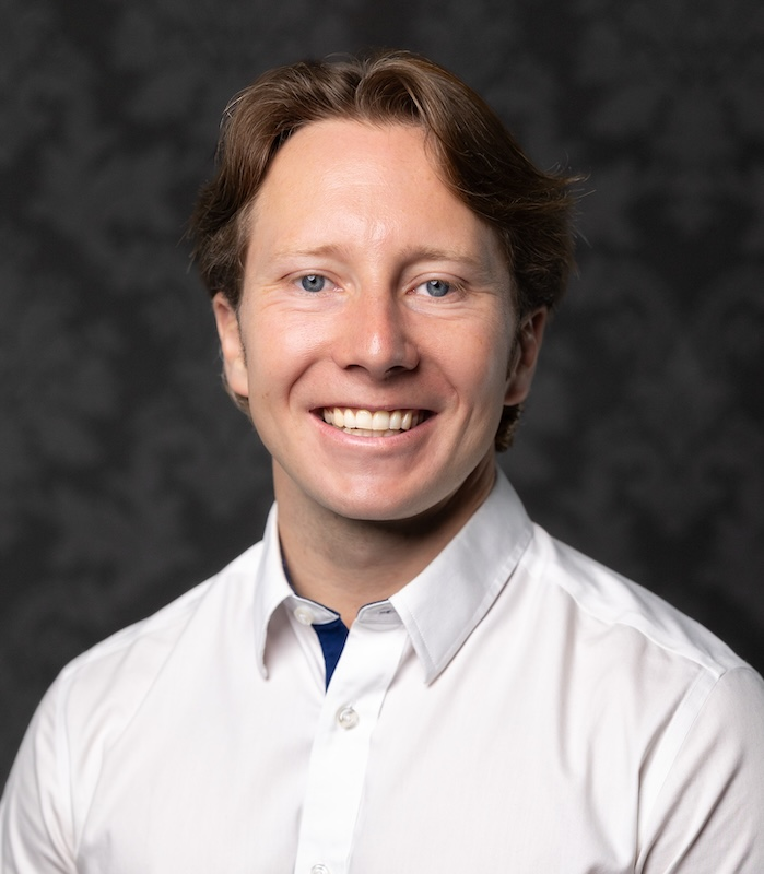 Man with light hair smiles wearing a white shirt; background is dark grey.