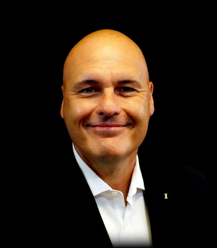 Bald man in a black suit smiles, white shirt open at the collar, against a black backdrop.