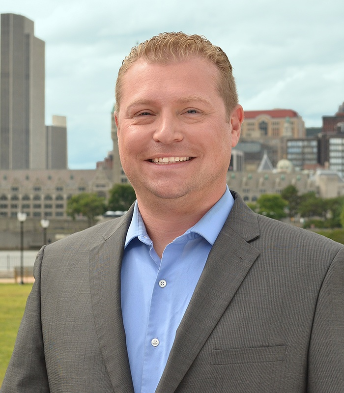 Man in a grey suit and blue shirt smiles, posing outdoors with city buildings in background.