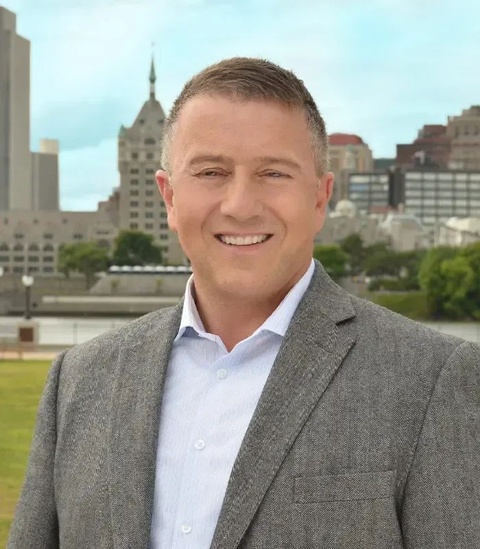 Man in blazer smiling outdoors, city skyline background.