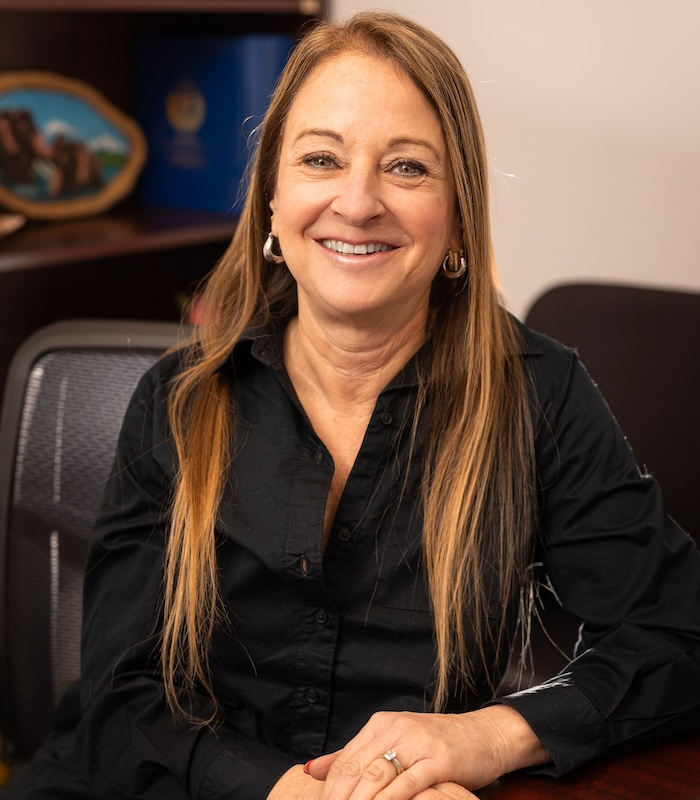 Woman with long blonde hair, smiling, wearing a black shirt, seated in an office.