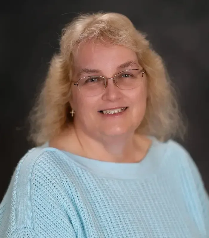 Woman with blonde hair, glasses, and blue sweater smiles.
