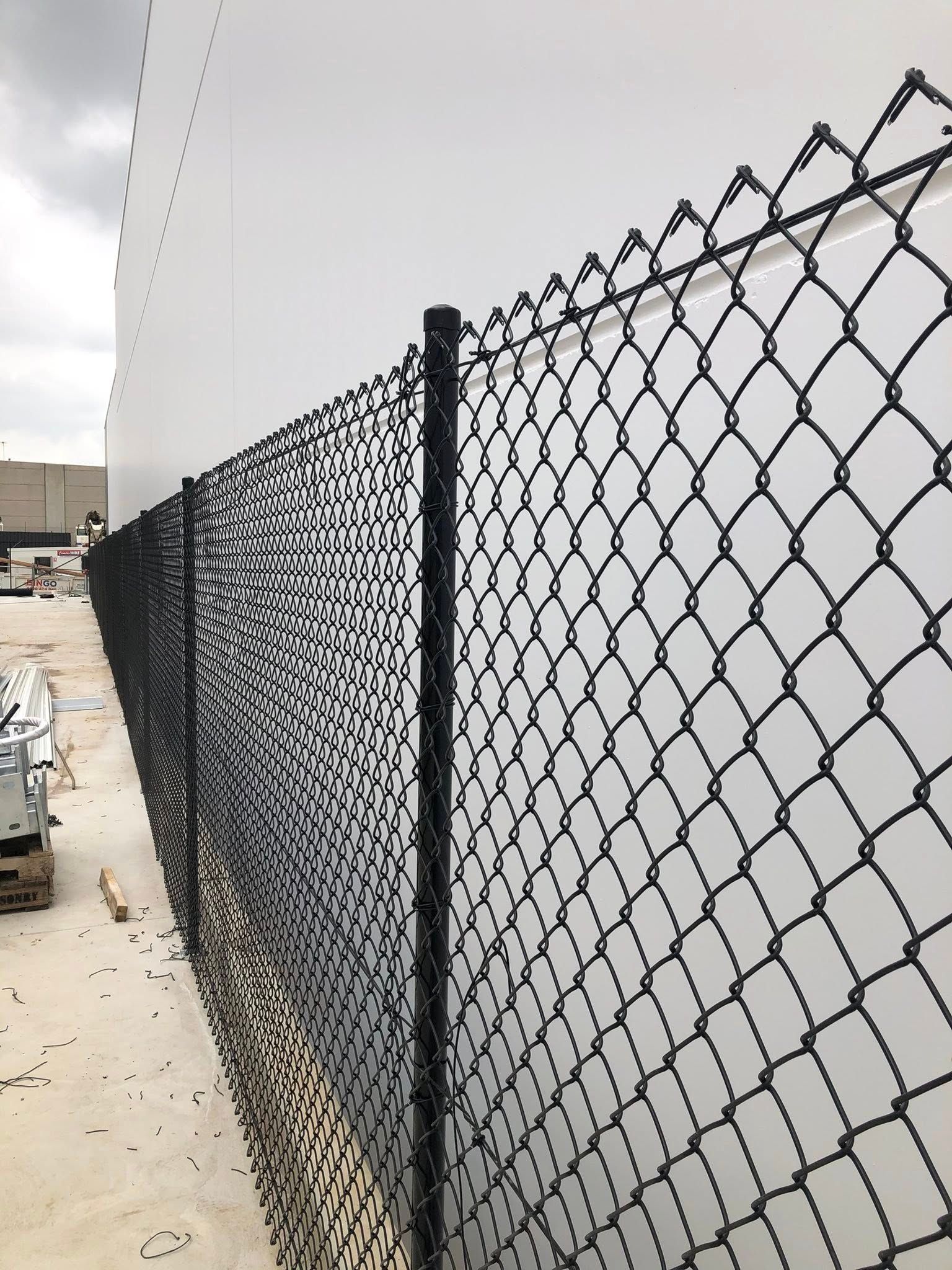 Black chain-link fence along a concrete path beside a large white wall under a cloudy sky.